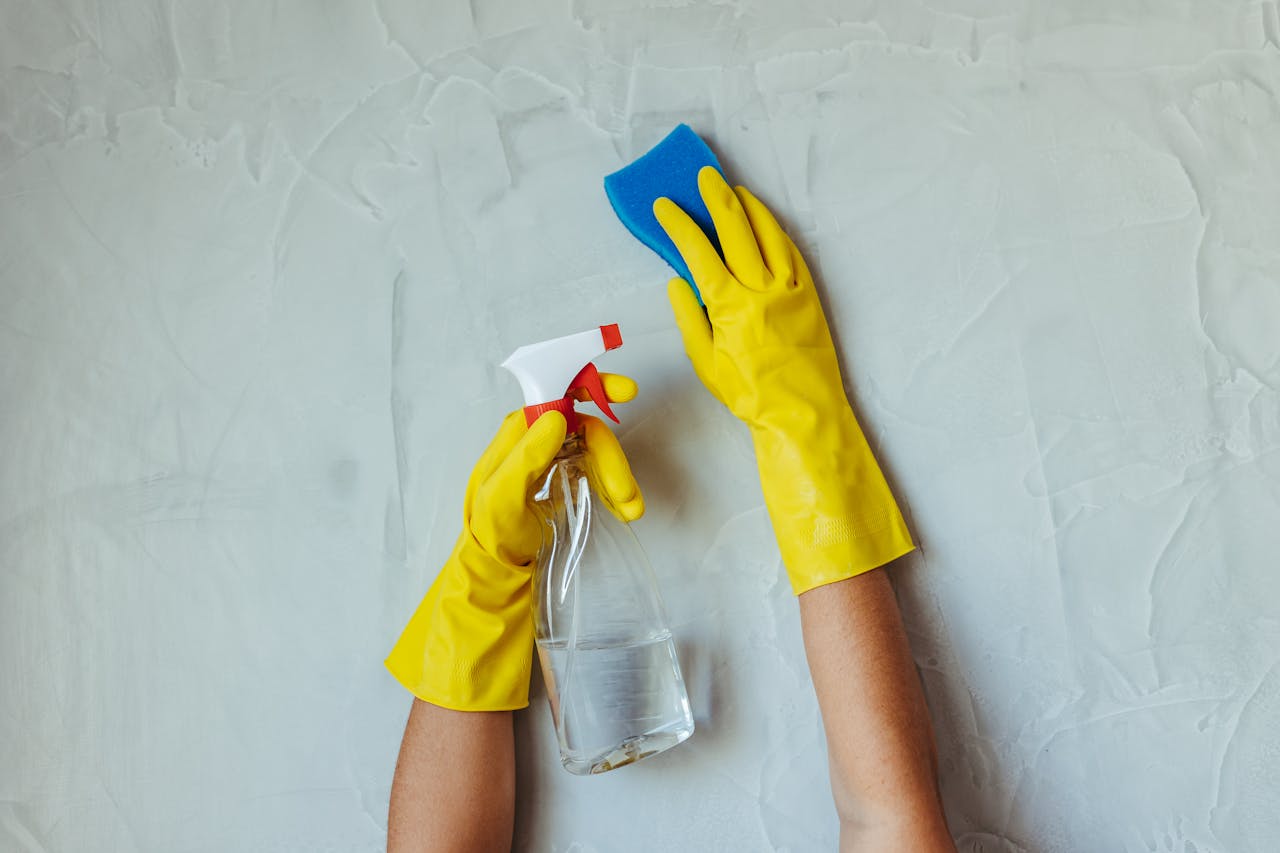 expertise-01 Close-up of hands in yellow gloves cleaning a wall with a spray bottle and sponge.